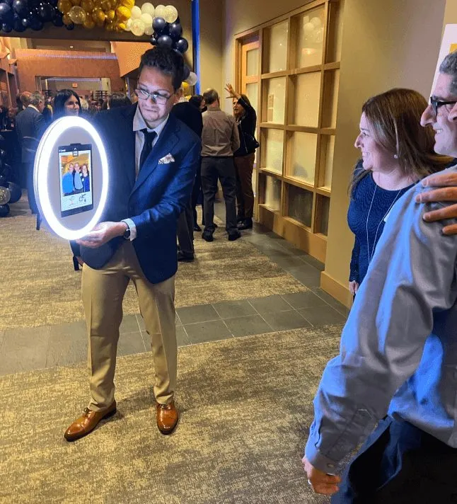 Attendant holding a handheld roaming photo booth with its ring light on at a party.